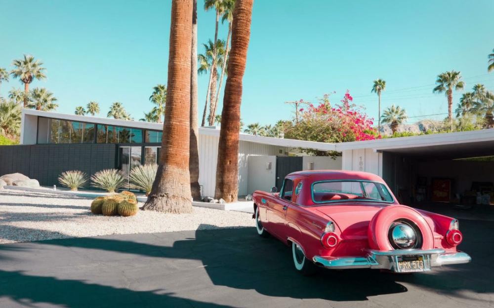 A vibrant red classic car parked in Palm Springs with mid-century modern architecture in the background.